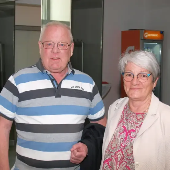 Ein älterer Mann und eine ältere Frau stehen in einer Innenumgebung nebeneinander. Der Mann trägt ein gestreiftes Hemd und die Frau trägt eine gemusterte Bluse und eine Brille. Ein Kühlschrank mit einem gelben Etikett ist im Hintergrund zu sehen.