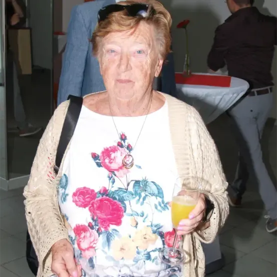 Eine ältere Frau mit einer Brille auf dem Kopf hält ein Glas Orangensaft in der Hand und steht in einem Raum mit einem Tisch dahinter.