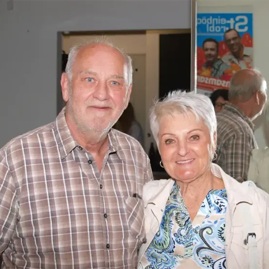 Ein älteres Paar steht gemeinsam in einem Raum und lächelt für ein Foto. Der Mann trägt ein kariertes Hemd, und die Frau hat kurze weiße Haare. Hinter ihnen ist ein Plakat mit Gesichtern zu sehen, und mehrere Personen sind im Hintergrund.