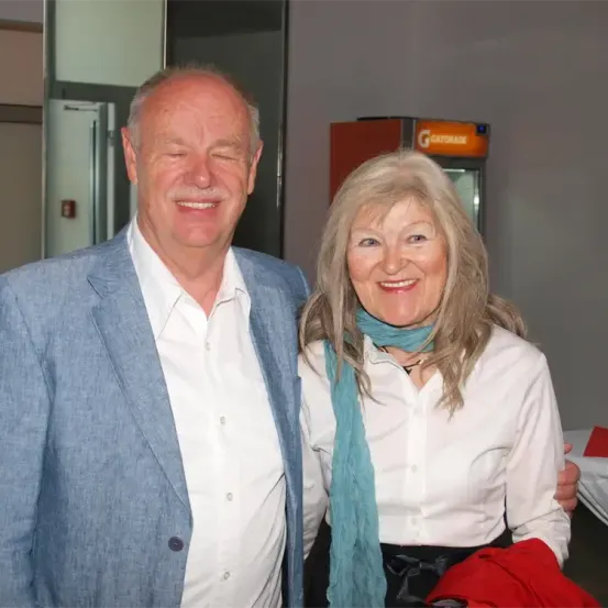 Ein älterer Mann und eine ältere Frau stehen lächelnd eng beieinander. Der Mann trägt einen blauen Anzug, die Frau eine weiße Bluse mit einem blauen Schal. Dahinter steht ein roter Kühlschrank mit dem Wort Gatorade.