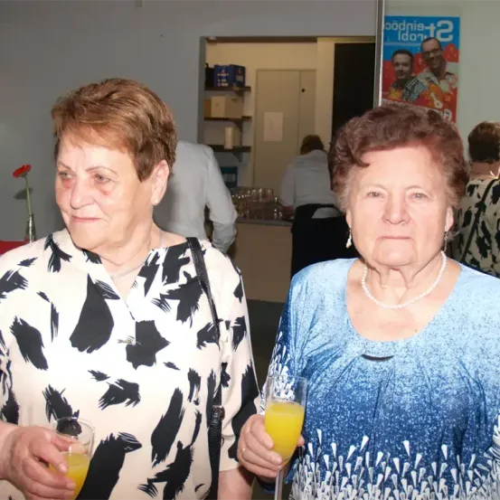 Zwei ältere Frauen, die Gläser mit Orangensaft halten, stehen in einem Raum mit einem Poster im Hintergrund.