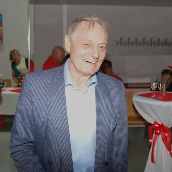 Ein älterer Mann mit einem blauen Anzug und einem blauen Hemd lächelt in einem Innenraum. Dahinter steht ein Tisch mit Weingläsern.