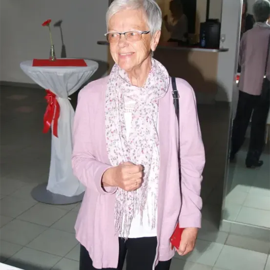 Eine ältere Frau mit Brille und einem Blumenschal steht lächelnd in einem Raum. Hinter ihr ist ein Tisch mit einem roten Band und Blumenzentrum zu sehen, und eine Person steht in der Nähe eines Spiegels.