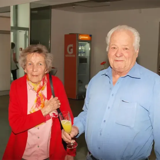 Ein älterer Mann und eine ältere Frau stehen nebeneinander und lächeln. Der Mann hält ein Glas Orangensaft. Hinter ihnen steht ein roter Kühlschrank mit einer Blume und einer Person im Hintergrund.