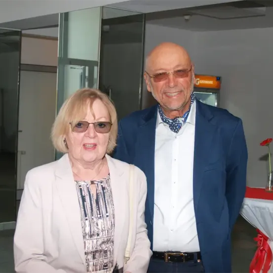 Ein älterer Mann und eine ältere Frau stehen nebeneinander in einem Raum, beide lächeln und tragen eine Brille. Der Mann trägt einen blauen Anzug mit Krawatte und die Frau eine weiße Jacke und Bluse. Hinter ihnen steht ein Tisch mit einem roten Tuch und einer Blume.