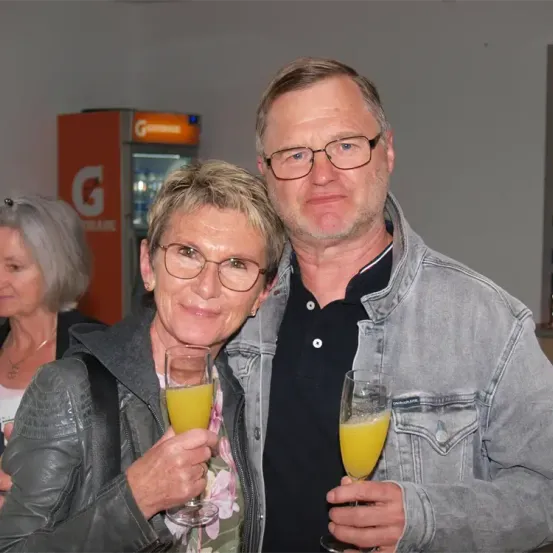 Ein älterer Mann und eine ältere Frau lächeln und halten Weingläser mit Orangensaft gefüllt, posieren für ein Foto. Hinter ihnen ist eine Frau mit grauen Haaren und ein orangefarbener Verkaufsautomat mit dem Buchstaben G darauf.