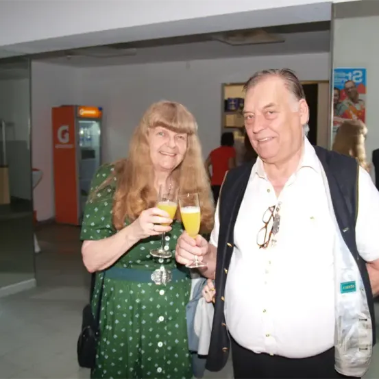 Ein lächelndes Paar in einem Raum, die Frau in einem grünen Kleid hält zwei Gläser Orangensaft, während der Mann in einem weißen Hemd lächelt und ein Glas hält. Hinter ihnen sind ein Kühlschrank und ein Poster zu sehen.
