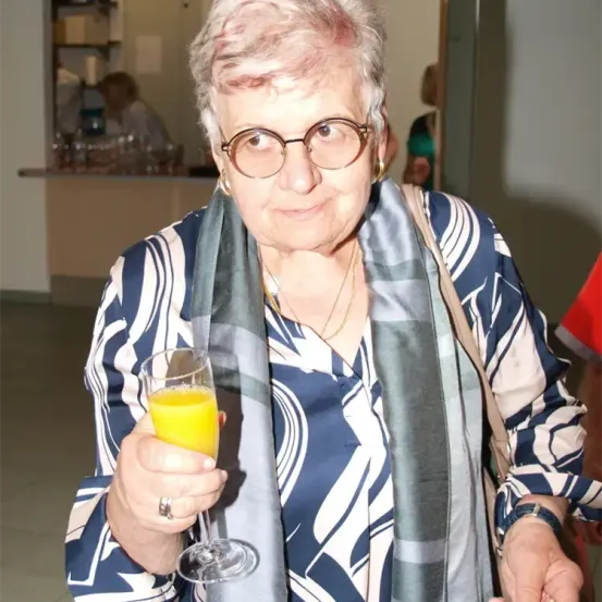 Eine ältere Frau mit Brille und Schal hält ein Glas Orangensaft. Sie steht drinnen, der verwackelte Hintergrund zeigt einen Tresen und Menschen.