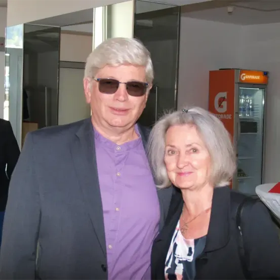 Ein älterer Mann und eine ältere Frau stehen zusammen und lächeln für ein Foto. Der Mann trägt ein lila Hemd und eine Sonnenbrille, während die Frau weißes Haar und eine Halskette hat. Hinter ihnen ist ein Verkaufsautomat und eine Glaswand zu sehen.