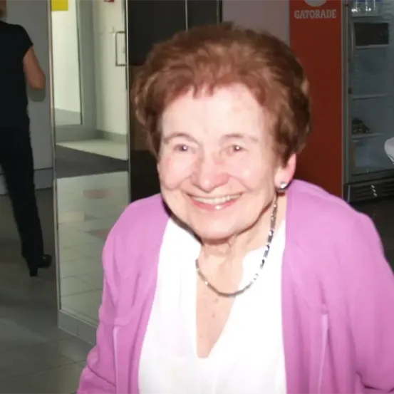 Eine ältere Frau mit roten Haaren lächelt in einem Flur. Sie trägt einen rosa Pullover und eine Halskette. Hinter ihr geht eine Person in einem Spiegel.