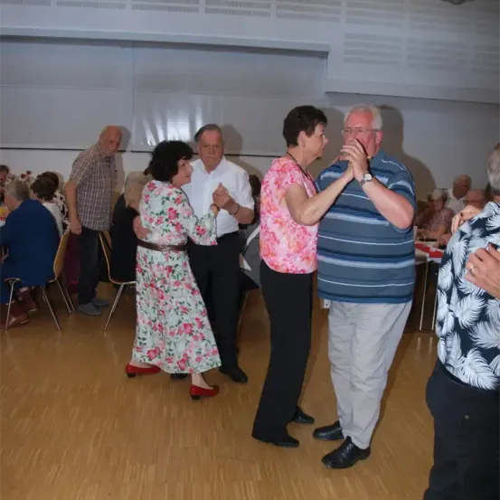 Eine Gruppe älterer Menschen tanzt in einem Raum. Sie lächeln und scheinen es zu genießen. Sie sind in Freizeitkleidung gekleidet. Hinter ihnen sitzen einige Leute auf Stühlen und andere stehen. Der Raum hat Holzböden und weiße Wände.