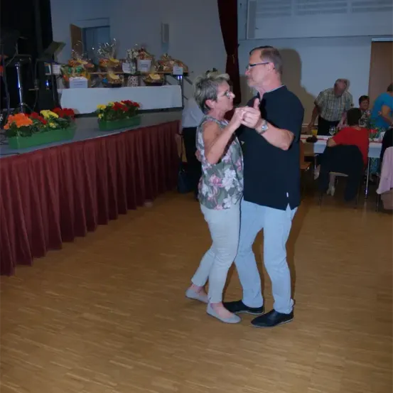 Eine Indoor-Tanzfläche, ein Paar tanzt mit einem Tisch mit Blumen im Hintergrund. Andere Leute sitzen an Tischen und essen möglicherweise.