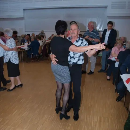 Ein Paar tanzt in der Mitte eines Raumes, umgeben von anderen, die zusehen und an Tischen sitzen. Der Raum hat Holzböden und weiße Wände.