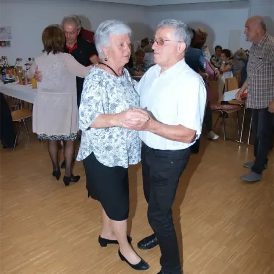 Ein älteres Paar tanzt in einer Halle, während andere sich unterhalten und ein Mann zuschaut. Ein Tisch mit Getränken und Blumen ist im Hintergrund.