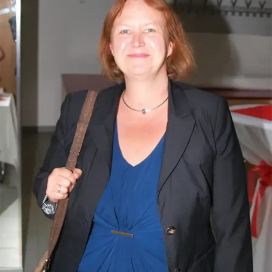 Eine Frau mit roten Haaren lächelt, trägt ein blaues Kleid und einen schwarzen Blazer. Sie hat eine Halskette und eine braune Tasche über der Schulter. Der Hintergrund ist ein Raum mit weißen Wänden und einer rot-weißen Tischdecke.