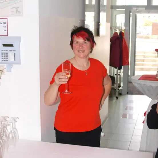 Eine Frau mit roten Haaren steht in einem Raum, hält ein Glas Wein in der Hand und lächelt. Sie trägt ein rotes Shirt und schwarze Hose. Eine weiße Wand mit einem Nummernschild und einem elektronischen Gerät befindet sich hinter ihr.
