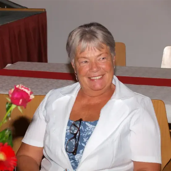 Eine ältere Frau mit grauem Haar lächelt und sitzt auf einem Stuhl. Sie trägt ein weißes Hemd und hat eine Brille auf der Brust.