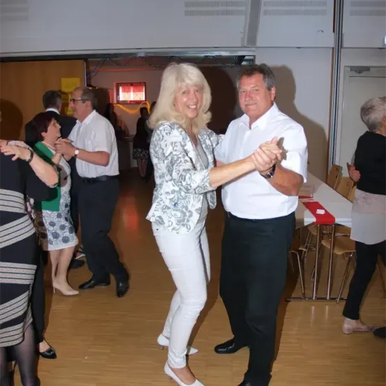 Eine Gruppe von Menschen tanzt in einem Saal. Eine Frau hält die Hand eines Mannes. Dahinter tanzen einige Leute. Tische und Stühle sind auf der rechten Seite angeordnet.