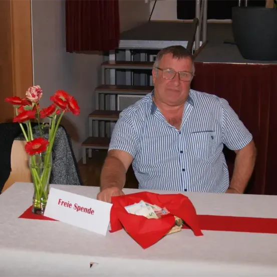 Ein Mann sitzt an einem Tisch mit einem Strauß roter Blumen und einem Schild, das 'Freie Spende' besagt. Dahinter befinden sich eine Treppe und ein Topf mit Pflanze. Er trägt eine Brille und ein gestreiftes Hemd.