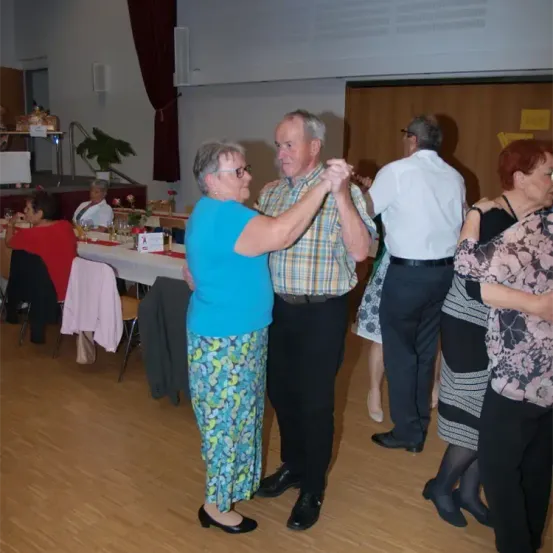Eine Gruppe älterer Erwachsener tanzt auf einem Holzboden. Eine Frau in Blau und ein Mann in einem karierten Hemd führen den Tanz an. Hinter ihnen stehen andere, darunter ein Mann in einem weißen Hemd und eine Frau in einem Blumenkleid. Im Hintergrund befindet sich ein Tisch mit Blumen und eine Bank mit Jacken.