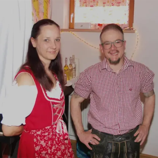 Ein Mann und eine Frau stehen nah beieinander in einem Raum und lächeln. Die Frau trägt ein traditionelles deutsches Kleid. Hinter ihnen stehen Flaschen und ein Regal.
