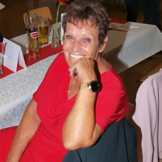 Eine ältere Frau in einem roten Kleid lächelt in die Kamera, sitzend an einem Tisch mit Biergläsern. Eine weiße Tischdecke, Namenskarten und eine Blumenvase sind auf dem Tisch.