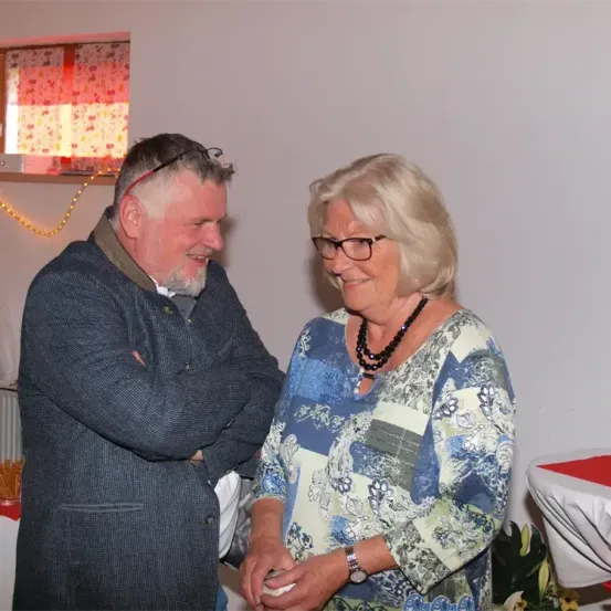 Ein älterer Mann und eine ältere Frau stehen nah beieinander und lächeln. Er trägt eine graue Jacke, sie ein blaues Blumenblusenkleid. Dahinter eine weiße Wand mit einem verhangenen Fenster.