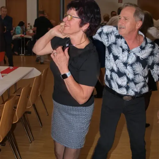 Eine Frau in einem schwarzen Oberteil und einer Rock tanzt mit einem Mann in einem schwarz-weißen Hemd. Sie befinden sich in einem Saal mit mehreren anderen Leuten, die sitzen und stehen.