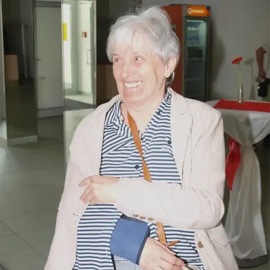 Eine ältere Frau mit weißen Haaren, die eine weiße Jacke und ein gestreiftes Shirt trägt, lächelt und posiert für ein Foto. Sie trägt eine blaue Tasche über der Schulter. Hinter ihr ist ein Tisch mit rotem Tuch und einer Blumenvase zu sehen. Ein Kühlschrank mit gelbem Aufkleber ist ebenfalls vorhanden.