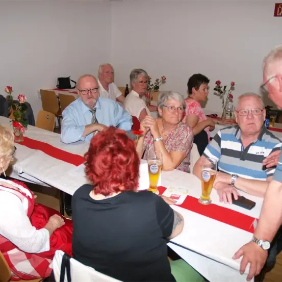 Mehrere ältere Personen sitzen in einem Esszimmer um einen Tisch herum und unterhalten sich vermutlich. Biergläser stehen auf dem Tisch, und eine Person hält ein Handy. Es gibt rote Rosen in Vasen auf dem Tisch und eine rot-weiße Tischdecke.
