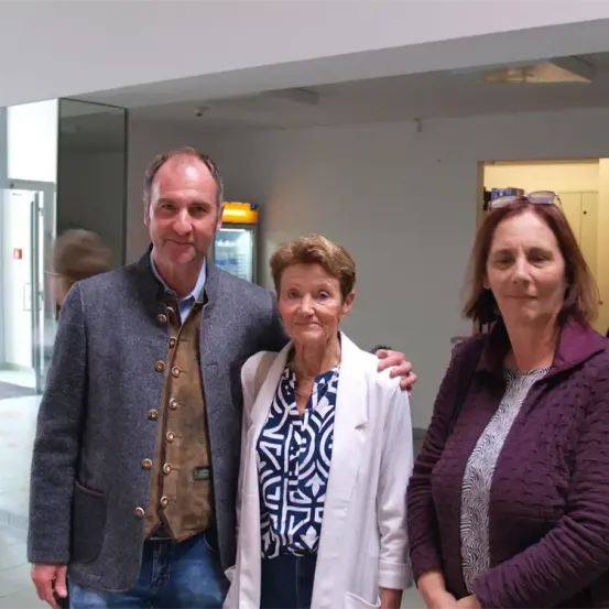 Drei Personen stehen in einem Flur, mit einem Mann links, einer Frau in der Mitte und einer Frau rechts. Der Mann trägt einen grauen Mantel, während die Frau in der Mitte einen weißen Blazer trägt. Die Frau rechts trägt eine lila Jacke. Sie scheinen für ein Foto zu posieren.