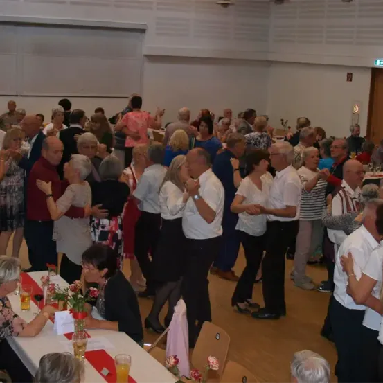 Ein Raum voller älterer Erwachsener versammelt, einige stehen, während andere um einen Tisch herum sitzen. Der Raum hat weiße Wände und Holzböden. Einige Menschen umarmen sich, und andere klatschen.