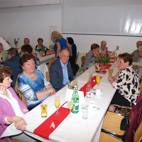 Eine Gruppe von Menschen sitzt an einem langen Tisch und lächelt. Auf dem Tisch stehen Blumen, Gläser und Teller. Sie scheinen eine Mahlzeit zu genießen.