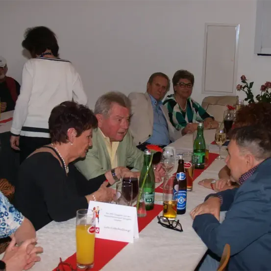 Eine Gruppe von Menschen sitzt an einem Tisch und unterhält sich. Flaschen und Gläser sind auf dem Tisch angeordnet, zusammen mit einigen Blumen. Eine Frau in einer weißen Bluse steht hinter ihnen, und ein Mann mit einer Brille sitzt in der Nähe.