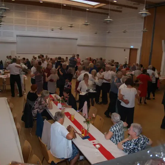 Eine Indoor-Veranstaltung mit älteren Menschen, einige tanzen und andere sitzen an Tischen mit Blumen und Gläsern. Die Tische sind mit rot-weißem Tuch bedeckt.