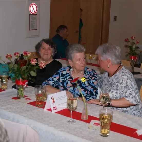 Drei ältere Frauen sitzen an einem Tisch mit Weinläsern, Blumen und einem Schild. Sie scheinen in ein Gespräch vertieft zu sein. Ein Schild mit Rauchverbot ist an der Wand zu sehen.