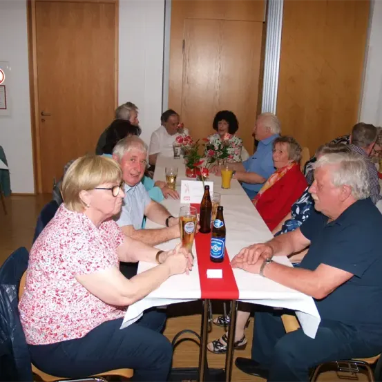 Eine Gruppe älterer Personen sitzt an einem langen Tisch und genießt Getränke und eine Blumenarrangement. Sie scheinen sich bei einem gesellschaftlichen Treffen zu befinden, vielleicht einer Wiedervereinigung.