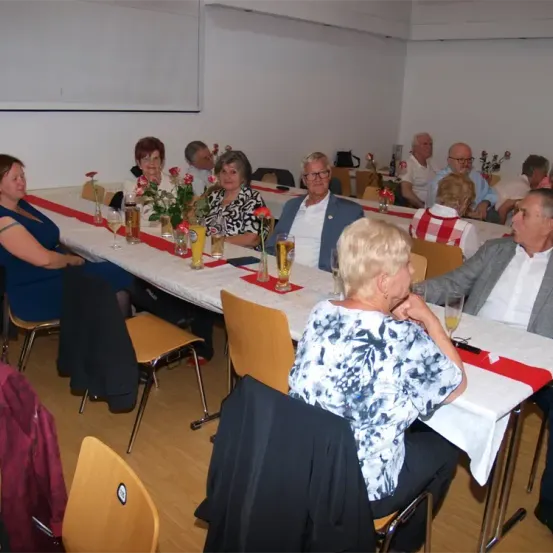 Eine Gruppe von Menschen sitzt an einem langen Tisch in einem Raum. Auf dem Tisch stehen Blumen und Gläser. Die Menschen tragen formelle Kleidung.