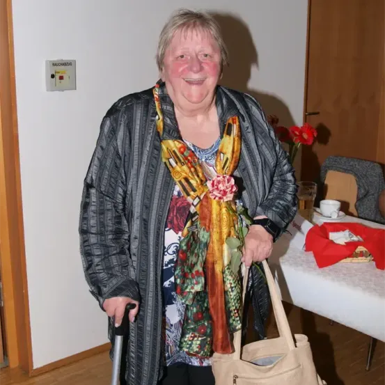 Eine lächelnde Frau mit Gehstock und buntem Schal steht neben einem Tisch mit Blumen und einer Tasse. Hinter ihr befindet sich ein Schild an der Wand.