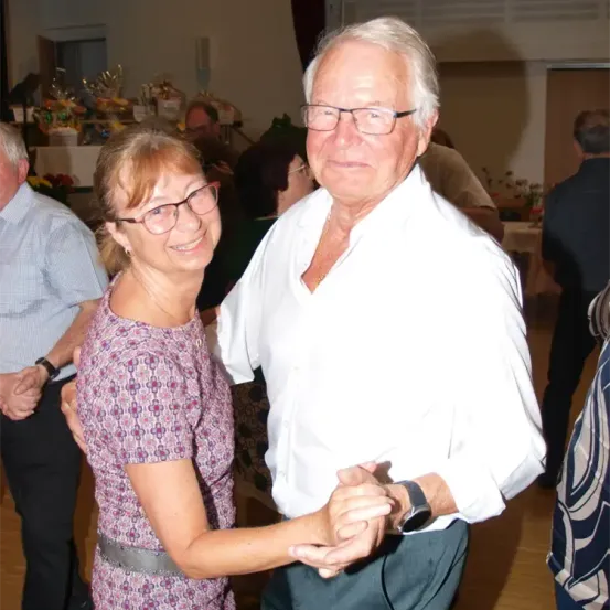 Ein älteres Paar tanzt bei einer Zusammenkunft im Innenbereich. Die Frau trägt eine Brille und ein Blumenkleid, während der Mann ein weißes Hemd und eine Brille trägt. Sie sind von anderen Menschen umgeben, einige stehend und einige sitzend, mit einem mit Blumen gefüllten Tisch im Hintergrund.