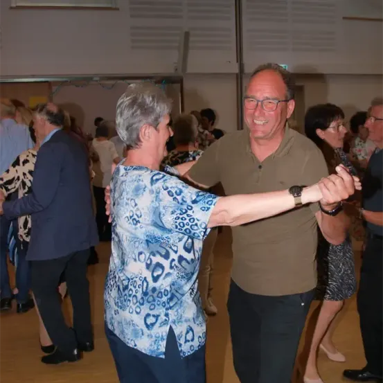 Ein Mann und eine Frau tanzen zusammen in einem Raum mit anderen Leuten. Sie lächeln und scheinen es zu genießen.