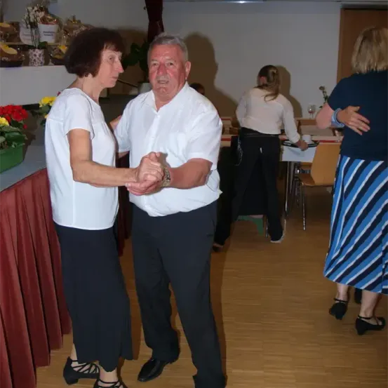 Ein Paar tanzt in einem Raum mit Holzböden und Tischen. Andere Menschen sind anwesend, darunter eine Frau in einem blauen Shirt und eine Frau in einem weißen Shirt.