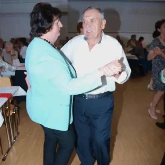 Ein älteres Paar tanzt in einem Raum mit Holzboden, umgeben von Menschen, die an Tischen sitzen. Die Frau trägt einen blauen Blazer, der Mann ein weißes Hemd und schwarze Hose.