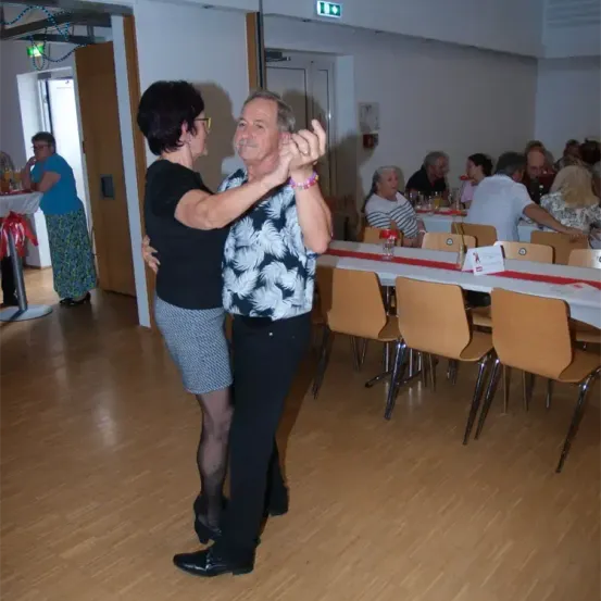 Ein älteres Paar tanzt auf einem Holzboden, während andere um Tische sitzen. Der Raum hat weiße Wände und einen Tisch mit roter Tischdecke.