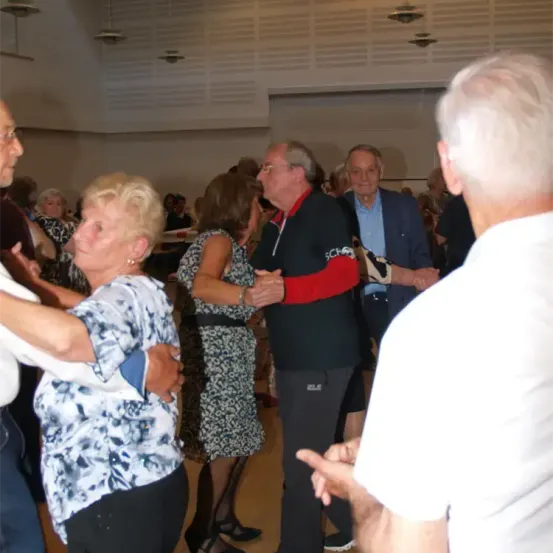Eine Gruppe älterer Menschen tanzt in einem großen Raum. Sie sind in Freizeitkleidung gekleidet. Der Mann in der Mitte trägt ein schwarzes Shirt mit rotem Kragen.