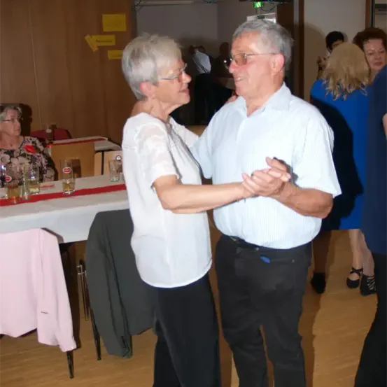 Ein älteres Paar tanzt in einem Raum mit Holzboden. Sie sind von anderen Menschen umgeben, die ebenfalls tanzen. Der Raum ist gut beleuchtet.