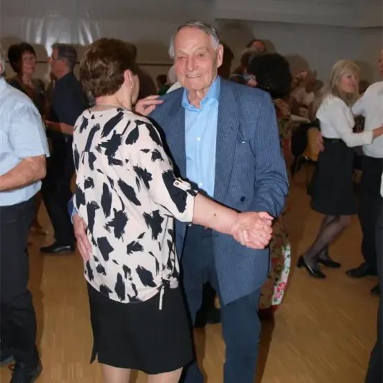 Ein älteres Paar tanzt in einer Halle mit anderen Paaren. Der Mann trägt einen marineblauen Anzug, und die Frau ein weißes und schwarzes Kleid. Sie sind von anderen Tänzern umgeben.