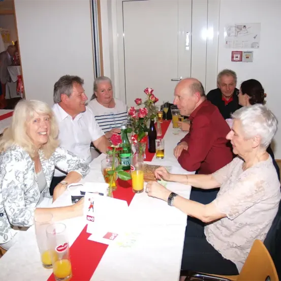 Eine Gruppe von Menschen sitzt um einen Tisch mit Blumen, Getränken und Karten. Sie scheinen sich bei einem geselligen Treffen zu amüsieren.