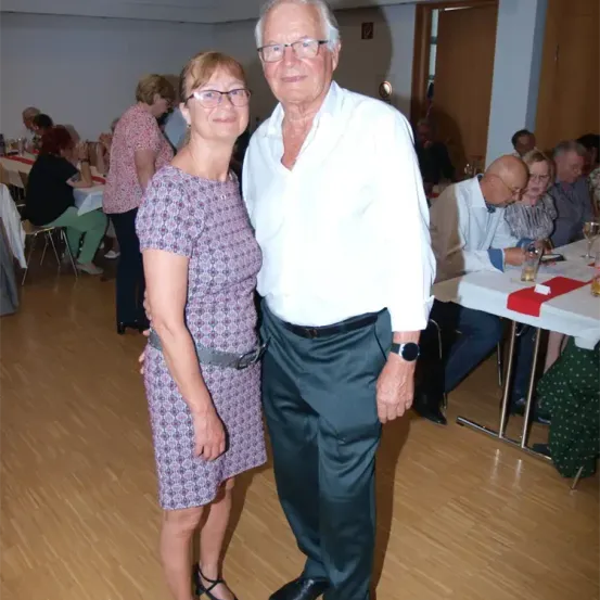 Ein älteres Paar steht in einem Speisesaal und lächelt. Hinter ihnen sitzen andere an Tischen und genießen eine Mahlzeit. Die Umgebung ist drinnen, mit Holzboden und weißen Wänden.