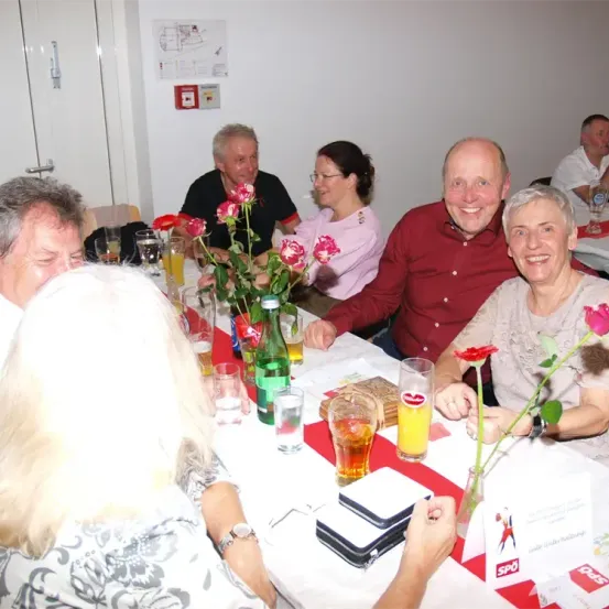 Mehrere Personen sitzen an einem langen Tisch mit Blumen und Getränken. Eine Frau in einem rosa Shirt sitzt in der Mitte, und ein Mann in einem roten Hemd lächelt. Dahinter sind eine Tür und ein Feuerlöscher zu sehen.